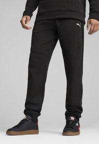 Svarta sweatpants med en åtsittande design, med en vit logotyp på vänster sida. Matchas med svarta sneakers med röda detaljer och en beige sula.