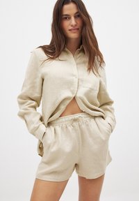 Beige linen button-up shirt with a chest pocket, paired with matching beige linen shorts that have an elastic waistband and drawstring.