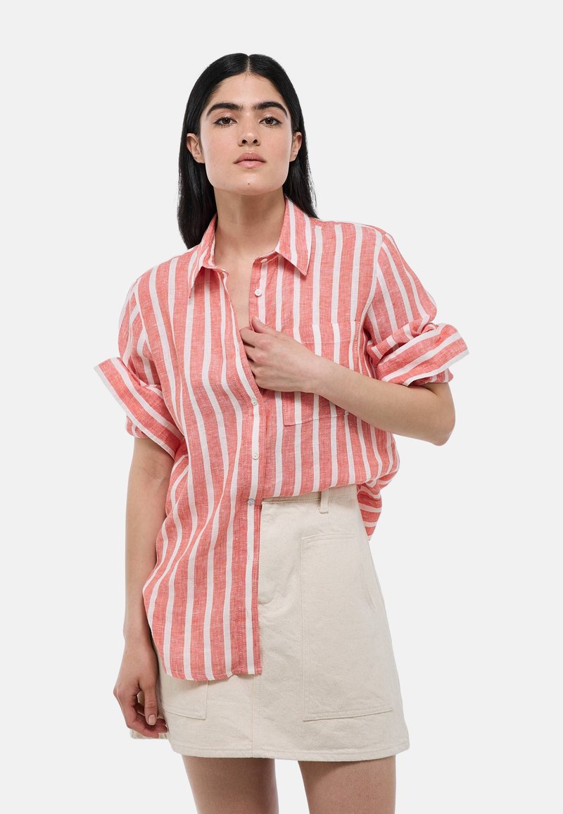 Chemise en lin rayée rouge et blanche à manches courtes et boutonnage à l'avant ; portée par-dessus une jupe beige clair dotée de poches à rabat.