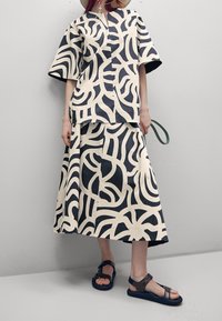 Set tunica e gonna a fantasia in crema e navy, con design astratti a onde. L'outfit è realizzato in un tessuto leggero, con maniche aperte. I sandali sono di un marrone scuro con un design a cinturino.
