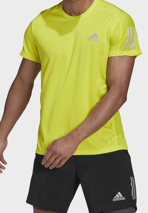 Homme portant un t-shirt Adidas jaune vif à manches courtes et un short Adidas noir avec logos, debout les bras légèrement pliés.