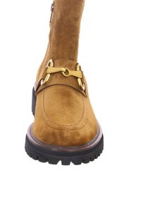 Botine din suede maro, cu vârf rotund, având accente de hardware aurii și o talpă groasă din cauciuc negru, cu cusături detaliate pe partea superioară.