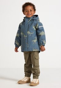Bambino sorridente con giacca blu stampata con dinosauri, pantaloni cargo color khaki e scarpe alte beige, in piedi su uno sfondo bianco uniforme.