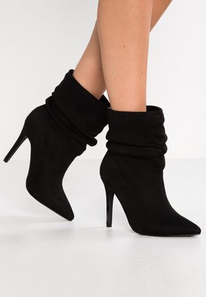 Jambes portant des bottines en daim noir décontractées, à bouts pointus et talons stiletto hauts, sur une surface blanche.