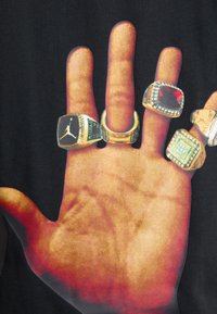 Černé tričko s velkým potiskem ruky, která zobrazuje pět prstenů. Prsteny jsou různých stylů, včetně čtvercových a kulatých tvarů, s kontrastními barvami.