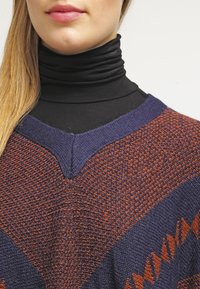 Kvinna som bär en svart polotröja under en texturerad navyblå och rostfärgad chevronmönstrad stickad tröja, med långt blont hår synligt.