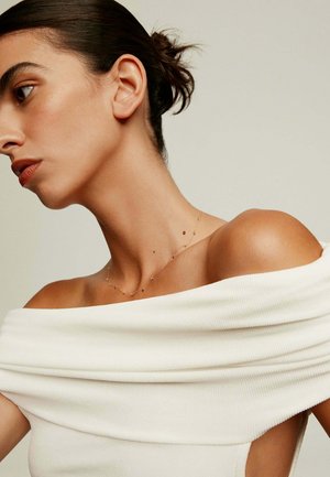 Top blanco de canalé con hombros al descubierto y cuello doblado, que muestra un delicado collar dorado con pequeños detalles en acento, con piel visible en el hombro.