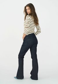 Camiseta de manga larga a rayas en color crema y marrón. Pantalones acampanados de mezclilla oscura con bolsillos. La modelo posa con las manos en las caderas, mirando ligeramente hacia atrás.