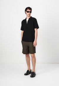 Polo scurt negru cu mânecă scurtă și detaliu cu fermoar, asociat cu pantaloni scurți verzi închis și mocasini negri. Material neted, croială modernă, design minimal.