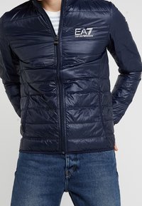 Námořnická pufovací bunda se zapínáním na zip, prošívaným designem a logem na hrudi. Vyrobena z lesklého materiálu, má stojáček a přiléhavé rukávy.