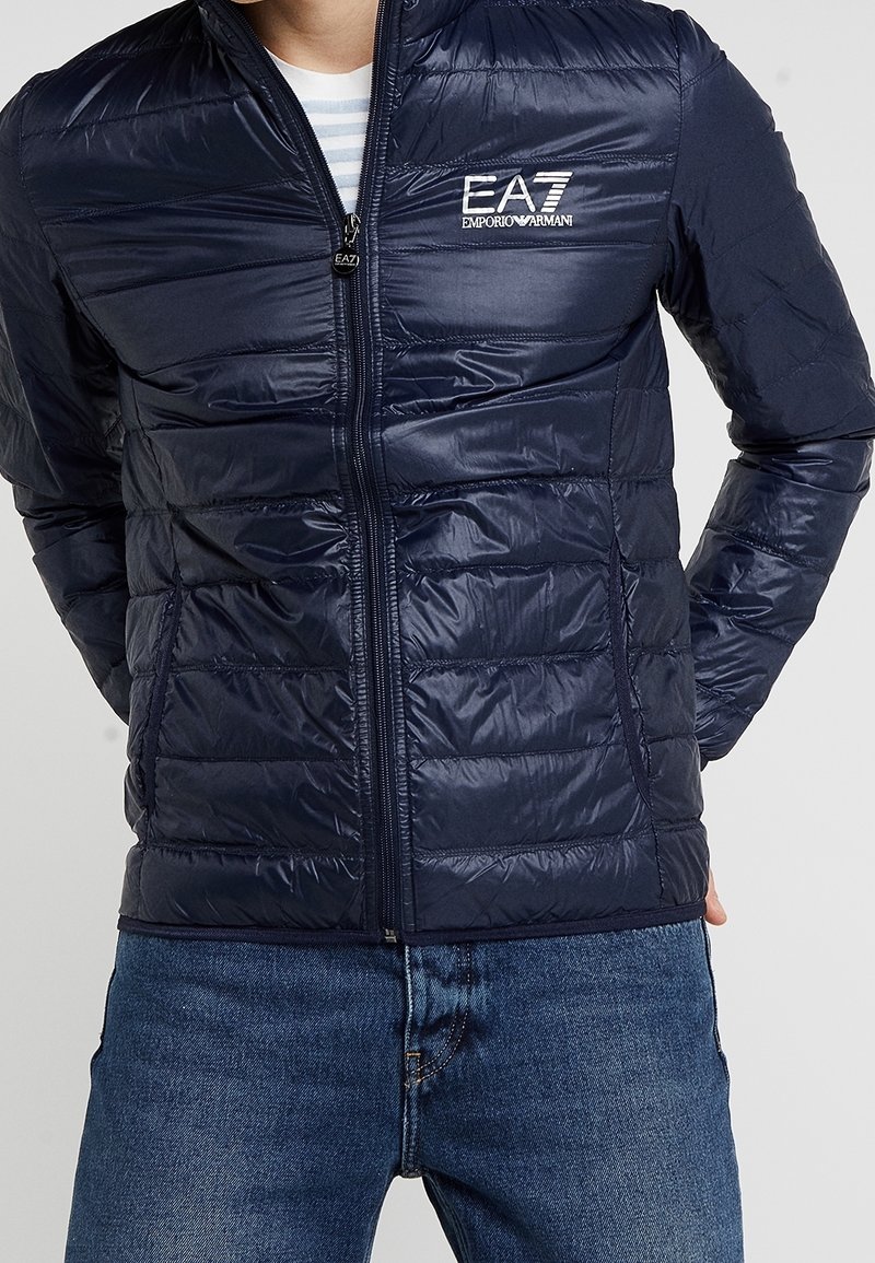 Námořnická pufovací bunda se zapínáním na zip, prošívaným designem a logem na hrudi. Vyrobena z lesklého materiálu, má stojáček a přiléhavé rukávy.