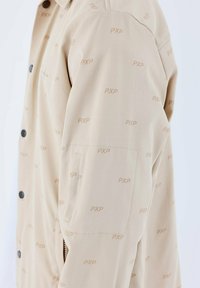 Chaqueta beige con un sutil patrón "PXP" en toda su superficie, con corte recto, cierre de botón y bolsillos laterales visibles. Textura mate.