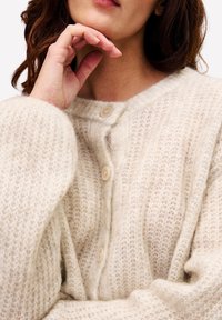 Vrouw in een crèmekleurige gebreide cardigan met knopen, die zachtjes haar kin aanraakt met haar hand bij haar gezicht.