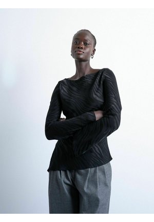 Femme debout, les bras croisés, portant un haut à manches longues noir à motifs et un pantalon gris, sur un fond blanc uni.