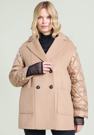 Woman with blonde hair wearing a beige coat featuring quilted sleeves and a double-breasted button front over a black top.