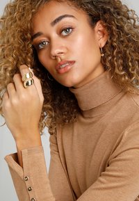 Beige turtleneck neule, jossa on ribbed kaula-aukko ja nappihihansuut; mallilla on päällä kultasävyinen kahta väriä sisältävä sormus ja renkaat, sekä lainehtivat hiukset.
