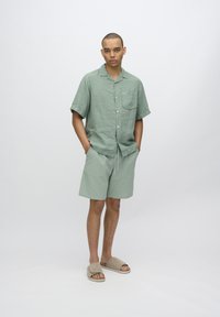 Light green linen short-sleeve shirt with a pocket, paired with matching patterned shorts and beige slide sandals, stands against a plain background.