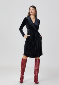Robe portefeuille en velours noir avec manches longues, ceinture et poches. Associée à des bottes hautes à motif serpent rouge. Design simple et élégant.