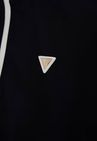 Crna tkanina s trokutastim bež kožnim zakrpa s reljefnim detaljima. Uključuje kontrastni bijeli patentni zatvarač.