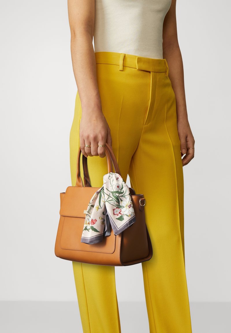 Femme tenant un sac à main en cuir beige avec une écharpe fleurie attachée à la poignée, portant un pantalon jaune et un haut blanc sans manches sur un fond uni.