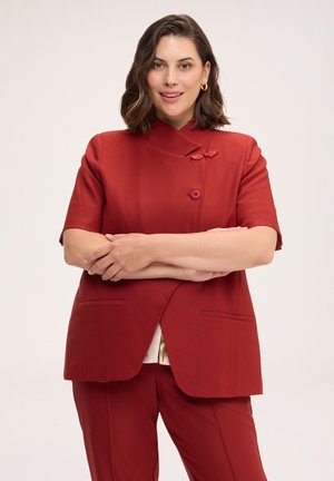 Vrouw met bruin schouderlang haar draagt een rode korte mouwen blazer en bijpassende broek, staat met gekruiste armen en glimlacht.