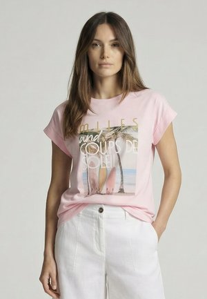 Woman with long brown hair wearing a pink T-shirt with palm tree and surfboard print, paired with white high-waisted pants, hand in pocket.