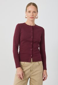 Burgundský cardigan s dlouhým rukávem vyrobený z měkké látky. Má kulatý výstřih a šest knoflíků vpředu, v kombinaci s béžovými kalhotami.