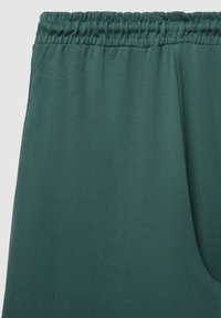 Grüne, elastische Sweatpants aus weichem Stoff. Sie verfügen über eine glatte Textur und seitliche Taschen mit einem lockeren Design. Keine sichtbaren Muster.