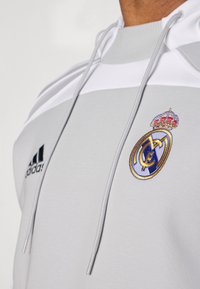 Sivi pulover dukserica s kapuljačom s bijelim detaljima, s vezanim logotipom Real Madrida u plavoj i zlatnoj boji, te logotipom Adidas na lijevoj strani.