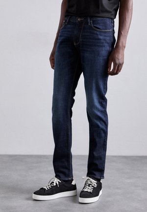 Homme portant un jean slim bleu foncé, des baskets noires avec semelles et lacets blancs, debout sur un sol gris devant un mur uni.