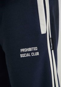 Marine sweatpants med hvide sidestriber. Har broderet tekst "PROHIBITED SOCIAL CLUB" i hvid. Blødt bomuldsstof, casual pasform.