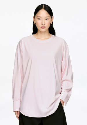 Light pink long-sleeve shirt with a round neck, dropped shoulders, and rounded hem. Fabric appears soft and slightly oversized.