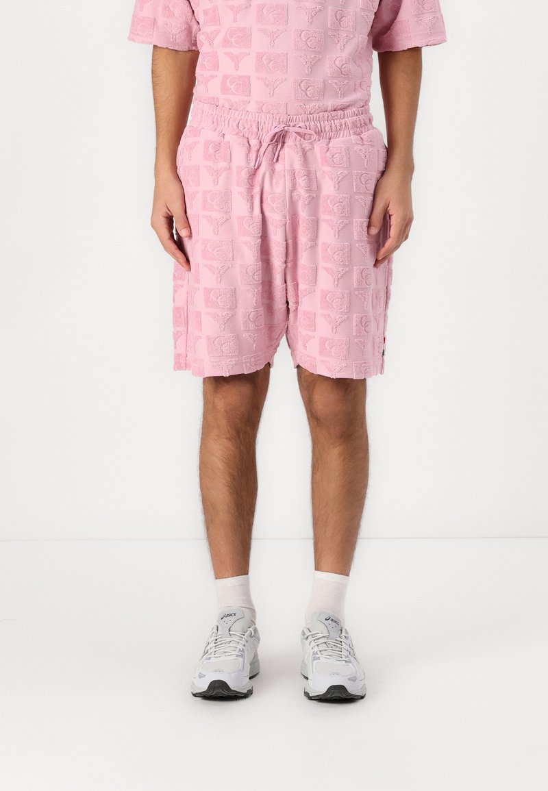 Pink textured shorts with elastic waistband and drawstring, featuring a repeated logo pattern. Worn with white ankle socks and sneakers.