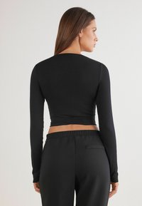 Černý dlouhý rukáv crop top z hladkého, pružného materiálu, v kombinaci s černými kalhotami s vysokým pasem, které mají nenápadný detail na zadní kapse.