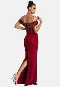 Rode avondjurk met een off-the-shoulder ontwerp, versierde lijfje, zijsplit en een slanke silhoutte. Heldere hakken.