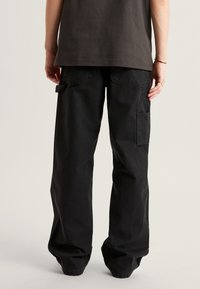 Pantalones cargo negros con un corte holgado, hechos de algodón resistente, que cuentan con bolsillos laterales y costuras robustas. La vista trasera muestra un diseño simple.