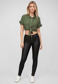 Cămașă scurtă de culoare verde măslinie, cu un nod în față, mâneci scurte și detalii cu butoane, asortată cu pantaloni skinny negri și adidași albi.