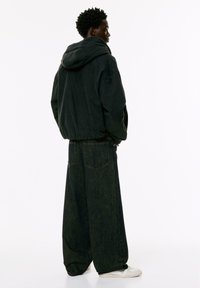 Felpa nera oversized con cappuccio regolabile, abbinata a jeans scuri a gamba larga. Accenti includono grandi tasche posteriori e sneakers bianche.