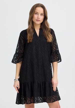 Žena s dlhými hnedými vlasmi, oblečená v čiernych priehľadných šatách so vzorom, s tromištvrťovými rozšírenými rukávmi a volánovou lemom, stojaca pred jednofarebným pozadím.