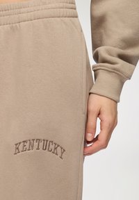 Pantaloni sportivi beige con vita elastica e tasca laterale. "KENTUCKY" è ricamato in un beige più scuro, curvato sull'orlo. Texture morbida.