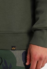 Felpa verde in cotone con polsini e orlo a costine, caratterizzata da un piccolo logo a patch nero e giallo vicino all'orlo. Pantaloni con motivo camouflage visibili sotto.