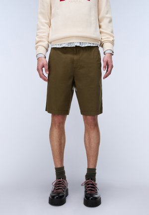 Mand iført olivengrønne knælange shorts, mørkegrønne sokker, sorte sko med røde snørebånd, cremefarvet sweater og en striped skjorte under.