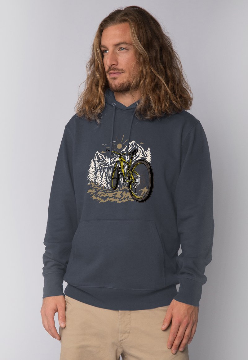 Mann mit welligem Haar, der einen marineblauen Hoodie trägt, der ein gelbes Mountainbike und weiße Kiefern mit Berg- und Sonnenhintergrund zeigt.