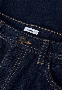 Tamsiai mėlyni džinsai iš džinso audinio su oranžinėmis siūlėmis, metaline sagtele užsegimui ir etikete su užrašu „TWO SOON“, dydis 28/32.