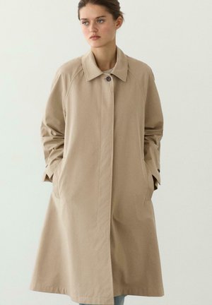 Frau trägt einen langen beigen Mantel mit einem Knopf und Kragen, die Hände in den Taschen, stehend vor einem einfarbigen hellen Hintergrund.