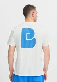 Baltos medvilnės marškinėliai su dideliu mėlynu stilizuotu 'B' grafiku nugaros pusėje. Trumpomis rankovėmis, apvaliu kaklu, laisvu siluetu.