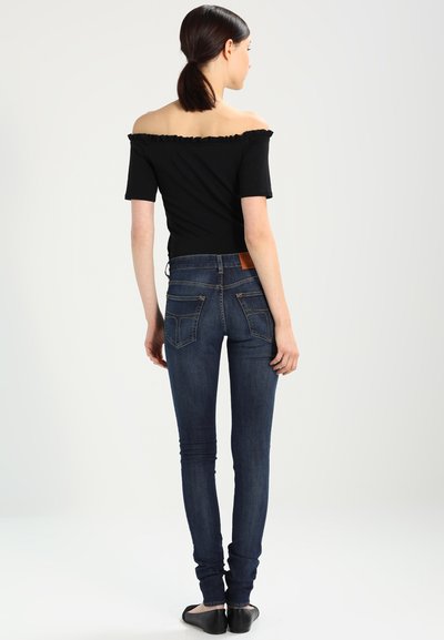 Een off-the-shoulder zwart top met een rimpelige halslijn, gecombineerd met slim-fit donkerblauwe jeans met subtiele stiksels en een bruine leren label op de achterkant.