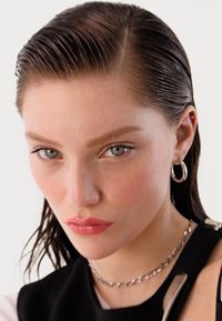 Close-up of a person with wet, slicked-back dark hair, wearing silver hoop earrings and a chain necklace, with a neutral makeup look.