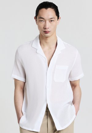 Junger Mann mit slickem Haar, der ein leichtes, kurzärmliges weißes Hemd und beige Hosen trägt und mit den Händen in den Taschen steht.