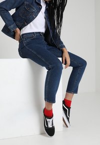 Jeansjacka, vit t-shirt, blå skinny jeans, svarta och vita sneakers, röda strumpor; sitter på en vit plattform mot en neutral bakgrund.